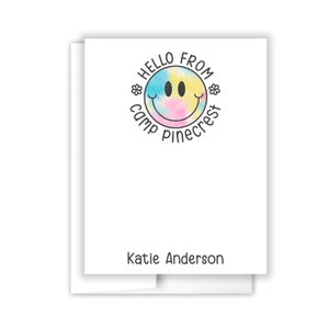 May include: A white card with a blue, yellow, and pink tie-dye smiley face with the text "Hello from Camp Pinecrest" around it. The name "Katie Anderson" is printed at the bottom of the card.