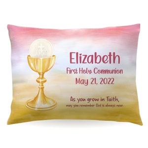May include: A pink and yellow pillow with a gold chalice and a white host with the word "IHS" on it. The text "Elizabeth First Holy Communion May 21, 2022 As you grow in Faith, may you remember God is always near." is printed on the pillow.