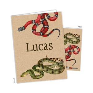 May include: A rectangular greeting card with a light brown kraft paper texture. The front features a red, black, and white coral snake, a green and black spotted snake, and the name Lucas in dark brown text. The back shows two snakes.