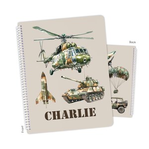 May include: Spiral-bound notebook featuring military vehicles: a helicopter, tank, rocket, and jeep with a parachute. The notebook has a tan cover with the name "CHARLIE" in bold letters. The back cover also has a military vehicle design.