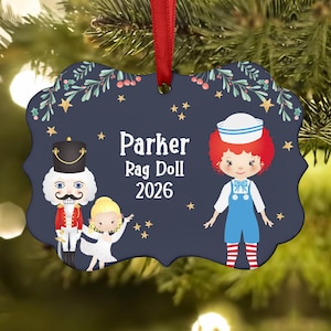 May include: A dark blue scalloped Christmas ornament featuring a nutcracker, a small child in a white dress, and a rag doll with red hair and a sailor hat. The text reads Parker, Rag Doll, 2026, surrounded by gold stars and green holly branches.