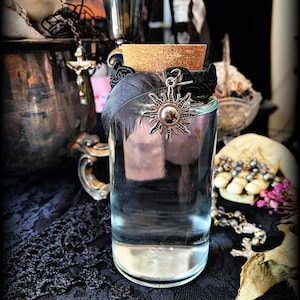 May include: A clear glass bottle with a cork stopper and a black ribbon tied around the top. A silver sun charm hangs from the ribbon. The bottle is sitting on a black lace tablecloth.