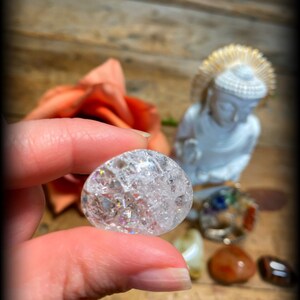 Pode incluir: Uma bola de cristal transparente e facetada com um efeito arco-&iacute;ris iridescente. A bola de cristal est&aacute; sendo segurada por uma m&atilde;o, com uma pequena est&aacute;tua de Buda e outros cristais no fundo.