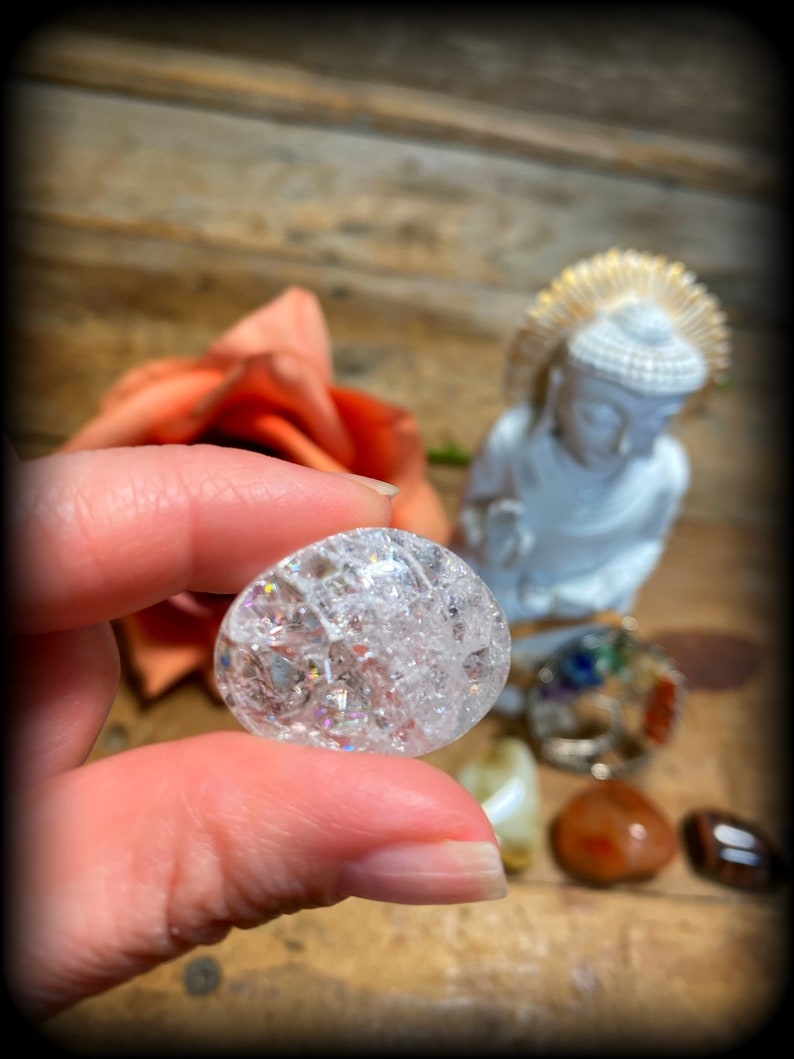 Pode incluir: Uma bola de cristal transparente e facetada com um efeito arco-&iacute;ris iridescente. A bola de cristal est&aacute; sendo segurada por uma m&atilde;o, com uma pequena est&aacute;tua de Buda e outros cristais no fundo.