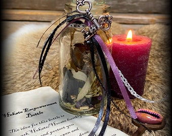 May include: A glass bottle with a key charm and purple ribbon, filled with dried herbs and flowers. The bottle is on a brown surface with a candle and a key with a seashell charm. The text on the paper reads "Hekate Empowerment Bottle. The idea for this bottle is to ask Hekate/Hecate humbly to guide and protect you with the bottle open and close it when you are done with your working or spell. Make sure to thank Her as you finish!"