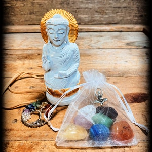 Pode incluir: Est&aacute;tua de Buda de madeira branca com uma aur&eacute;ola dourada, sentada sobre uma superf&iacute;cie de madeira. Em primeiro plano, uma pequena bolsa de organza branca com um amuleto prateado e sete pedras de chakra coloridas.
