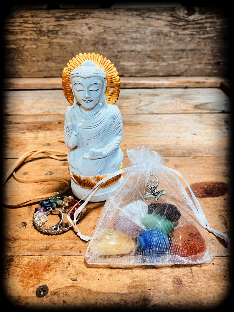 Pode incluir: Est&aacute;tua de Buda de madeira branca com uma aur&eacute;ola dourada, sentada sobre uma superf&iacute;cie de madeira. Em primeiro plano, uma pequena bolsa de organza branca com um amuleto prateado e sete pedras de chakra coloridas.