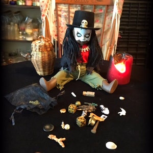 May include: A voodoo doll with long black hair, wearing a red shirt, black pants, and a black top hat with a skull on it. The doll is sitting on a black surface with a variety of objects scattered around it, including dice, a small bag, a candle, and a wooden stick.