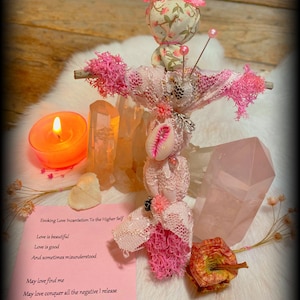 May include: A pink fabric doll with a white lace dress and a pink bow on its head. The doll is surrounded by crystals and a lit candle. The doll is holding a seashell. The text on the card reads: "Evoking Love Incantation To the Higher Self Love is beautiful Love is good And sometimes misunderstood May love find me May love conquer all the negative I release May it soothe me peace What I send out"