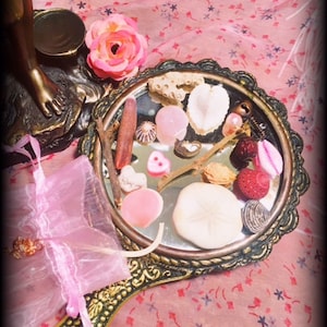 May include: A vintage hand mirror with a gold frame and a pink and white floral design. The mirror is filled with various small objects, including seashells, beads, and a pink heart.