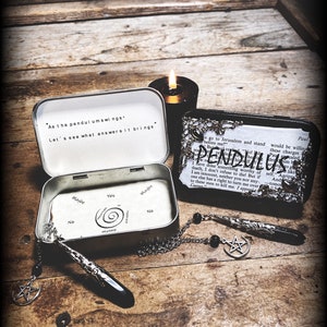 May include: A silver and black pendulum kit with a quote that reads "As the pendulum swings, Let's see what answers it brings." The kit includes a pendulum, a chain, and a small tin box with a chart inside. The box is open and the chart is visible. The chart has the words "Yes", "Maybe", "No", and "Maybe" written on it. The pendulum is hanging from the chain and is over the chart. There is a black candle in the background.