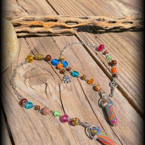 May include: A colorful beaded necklace with a peace sign charm and two rainbow-colored crystal pendants. The necklace is made with a variety of beads, including wood, glass, and metal.