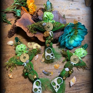 May include: Three green and gold decorative wands with a crystal point, seashells, and coins. The wands are adorned with green moss, netting, and beads. The wands are displayed on a wooden surface.