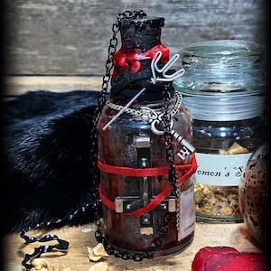 May include: A glass bottle with a black top and a red wax seal. The bottle is decorated with a black chain, a silver cross, and a red ribbon. The bottle is sitting on a wooden surface with some small pieces of dried fruit.