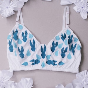 Puede incluir: Sujetador blanco con tirantes ajustables con un estampado azul y azul claro de gotas de lluvia y conejos estilizados.