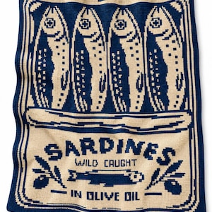 May include: A rectangular knit blanket featuring a dark blue and cream color scheme. The design depicts four fish above text that reads SARDINES WILD CAUGHT IN OLIVE OIL, flanked by two olive branches, all enclosed within a decorative border.