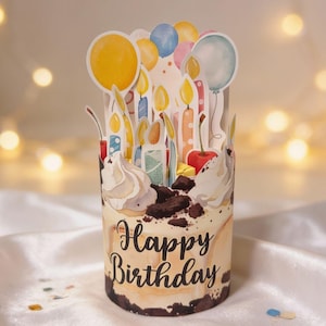 以下が含まれることがあります： 水彩デザインのケーキ型デコレーションバースデーカード。クリーム色のベースにチョコレートクランブル、ホイップクリーム、「Happy Birthday」のメッセージ。ろうそくと風船の切り抜きで飾られています。