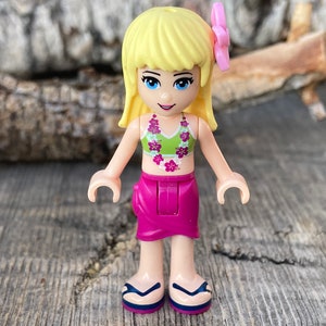 May include: A blonde Lego minifigure wearing a green and pink floral bikini top, a pink skirt, and a pink flower in her hair. She is wearing sandals and has a necklace with pink flowers.