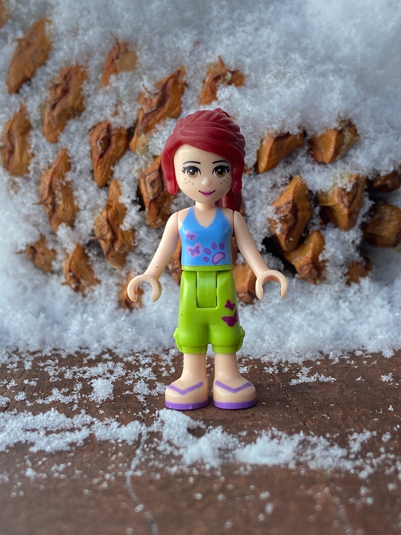Puede incluir: Una mu&ntilde;eca de pelo rojo con un atuendo azul y verde y sandalias moradas. La mu&ntilde;eca est&aacute; de pie sobre una superficie marr&oacute;n cubierta de nieve.