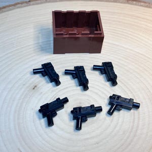 May include: Six black plastic toy guns, resembling machine guns, are arranged on a wooden surface. A brown plastic box is in the background.