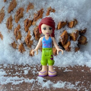 Puede incluir: Una mu&ntilde;eca Lego de pelo rojo que lleva una camiseta azul y morada, pantalones cortos verdes y sandalias moradas. La mu&ntilde;eca est&aacute; de pie frente a una pi&ntilde;a marr&oacute;n cubierta de nieve.
