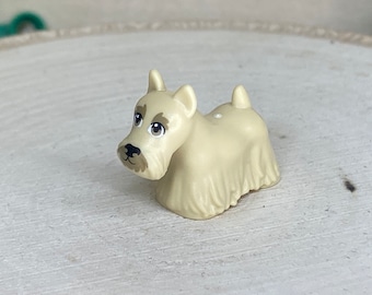 May include: A small, cream-colored figurine of a Scottish Terrier dog. The dog has a detailed face with brown eyes and a black nose. Its fur is textured, and it has a small tail. The figurine is set against a light-colored wooden surface.