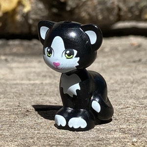 May include: A black and white cat figurine with green eyes and a pink nose. The cat is sitting with its paws tucked under its body.