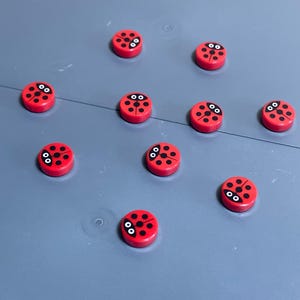 May include: A collection of red, circular ladybug-themed objects with black spots and white eyes. The objects are scattered on a gray surface. The ladybug design is consistent across all pieces, creating a playful and whimsical aesthetic.