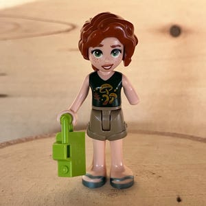 May include: A Lego minifigure of a girl with red curly hair wearing a green tank top with a mushroom design and brown shorts. She is holding a green flashlight.