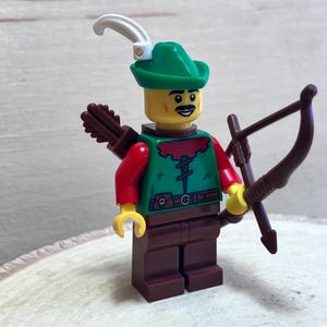 May include: A green and red Lego minifigure of a Robin Hood character with a brown bow and arrow, a green hat with a white feather, and a brown belt.