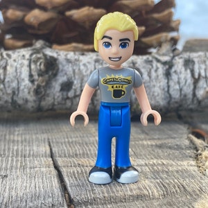 Puede incluir: Una minifigura de Lego de un hombre con cabello rubio, ojos azules y una expresión sonriente. Viste una camiseta gris con el texto "CAPES & COWLS CAFE" y un gráfico de taza de café, pantalones azules y zapatos grises y blancos. La figura está sobre una superficie de madera.