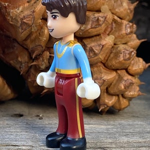 May include: A small plastic figurine of a prince wearing a blue shirt with gold trim, burgundy pants with gold stripes, and black shoes. The figurine has brown hair and a friendly expression.