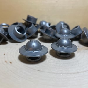 Puede incluir: Una colección de pequeños objetos plateados con forma de sombrero. Cada sombrero tiene un ala ancha y una parte superior redondeada con pequeños remaches decorativos. Los objetos están dispersos sobre una superficie de madera clara.