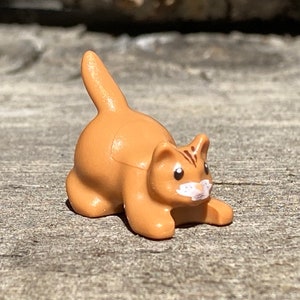 May include: A small, tan-colored cat figurine with a curved tail and white markings on its face. The cat is in a playful pose, with its front paws extended. The figurine is made of plastic and is a miniature collectible.