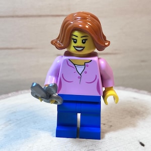 May include: A Lego minifigure of a woman with brown hair, wearing a pink shirt, blue jeans, and holding a pair of silver scissors.