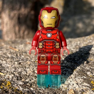 Puede incluir: Una minifigura Lego de Iron Man de color rojo y dorado. La figura lleva un traje rojo y dorado y está sobre una base de plástico transparente.