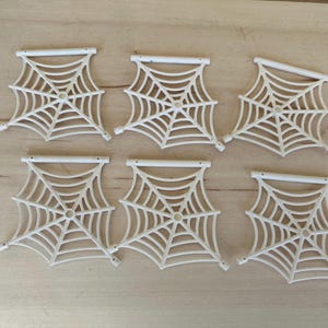 May include: Six white plastic spider web decorations. Each web is a flat, square shape with a raised center and a white plastic bar across the top.