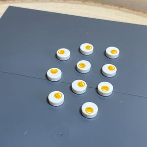 May include: A collection of eleven white, circular, decorative buttons, each featuring a yellow yolk design. The buttons are arranged on a gray surface, suggesting a craft or DIY project. The buttons are small, likely less than 1 inch in diameter.