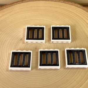 May include: Five white Lego window pieces with black bars are arranged on a light brown wooden surface. The windows have a rectangular shape with three vertical black bars. The pieces are designed for building and construction.