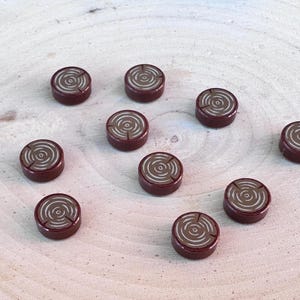 May include: A collection of ten small, round, dark brown buttons. Each button features a light-colored, engraved graphic design on the top surface that mimics the concentric rings found in a cross-section of a tree trunk or wood grain.
