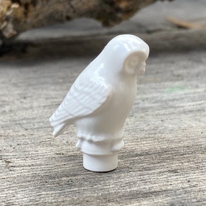 以下が含まれることがあります： 小さな白いセラミック製のフクロウの置物。フクロウは直立しており、翼と羽のテクスチャが詳細に表現されています。置物は滑らかで光沢のある仕上がりで、円筒形の台座に取り付けられています。背景は風化した木製の表面です。