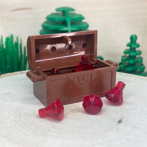 Puede incluir: Un cofre del tesoro de madera marrón con una tapa con bisagras está abierto, revelando varias gemas de plástico rojo en su interior. Cuatro gemas rojas adicionales están dispersas alrededor del cofre sobre una superficie de madera.