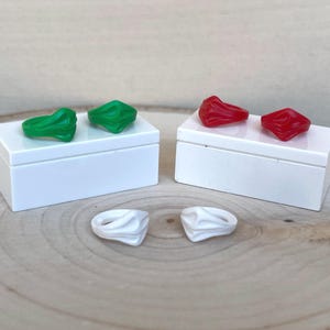 May include: A selection of rings in different colours. Two green rings and two red rings are on white boxes. Two white rings are on a wooden surface. The rings have a unique, textured design.
