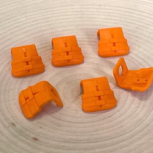 May include: Orange miniature life vests, a set of six, are arranged on a light wood surface. The vests have a detailed design with straps and buckles, and are brightly colored. One vest is open, showing the interior.