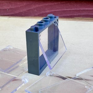 Może przedstawiać: Ciemnoniebieska cegła Lego z przezroczystym plastikowym oknem i kilkoma przezroczystymi plastikowymi panelami. Cegła Lego ma trzy okrągłe kołki na górze. Przezroczyste panele z tworzywa sztucznego są prostokątne z zaokrąglonymi narożnikami.