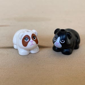May include: Two small, plastic animal figurines. One is white with brown markings around the eyes and ears, and the other is black with white and pink facial details. Both have a hole on top.