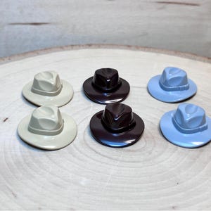 Op de afbeelding: Zes miniatuur vilt hoeden in verschillende kleuren, waaronder lichtblauw, donkerblauw, bruin en beige. De hoeden zijn in een cirkel op een houten oppervlak gerangschikt.
