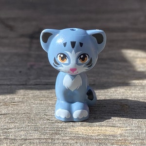 May include: A small blue and white plastic toy cat figurine with black ears and a pink nose. The cat has black stripes on its face and brown eyes.
