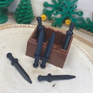 Puede incluir: Una colección de espadas de juguete negras, algunas dentro de una caja marrón, otras esparcidas sobre una superficie de madera. Las espadas tienen empuñaduras detalladas y hojas afiladas, que se asemejan a armas en miniatura. La escena se desarrolla sobre un fondo de elementos decorativos verdes.