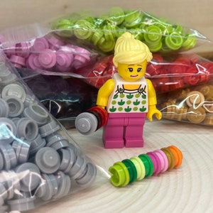 May include: A small plastic figurine of a person wearing a pink shirt with green apples on it. The figurine is surrounded by bags of small plastic pieces in various colors, including gray, pink, green, red, and gold.